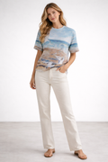 Woman wearing a patterned top with a beach and seagulls and white pants on a plain background